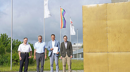 Vier Männer stehen neben einem großen goldenen Würfel, einem Kunstwerk am Bau, vor wehenden Flaggen, darunter die Regenbogenflagge und halten ein Blatt Papier hoch - lächelnd.