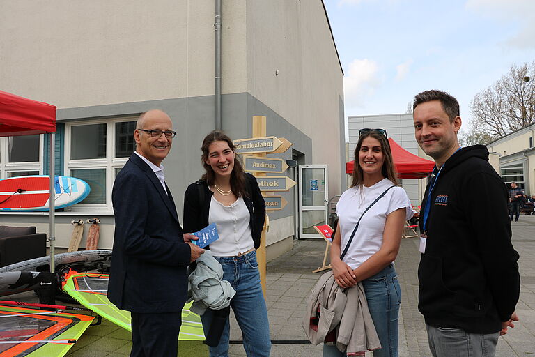 Zwei junge Frauen, ein Mann, Prof. Auerbach, und ein junger Mann , der wissenschaftliche Mitarbeiter benjamin Clausius im Gespräch vor einem Wegweiser, zu dessen Fuß SegelEquipment liegt 