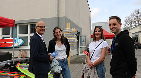 Zwei junge Frauen, ein Mann, Prof. Auerbach, und ein junger Mann , der wissenschaftliche Mitarbeiter benjamin Clausius im Gespräch vor einem Wegweiser, zu dessen Fuß SegelEquipment liegt 