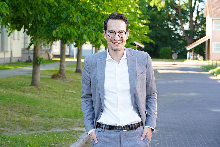 Ein lächelnder Mann im Anzug steht vor einer Baum-Allee auf dem Campus: Kanzler Fabian Berger