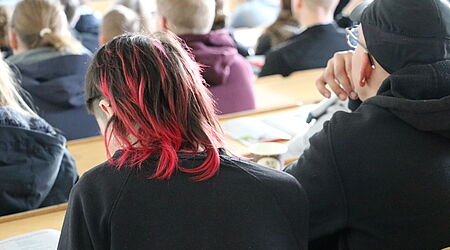 Eine Gruppe von vermeintlich jungen Menschen ist in einem Hörsaal zu sehen, eine Ansicht der Hinterköpfe.