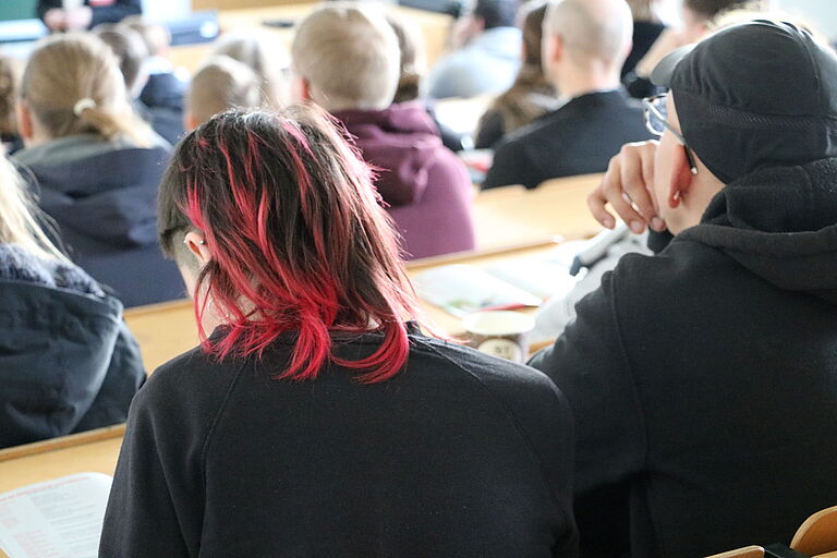 Eine Gruppe von vermeintlich jungen Menschen ist in einem Hörsaal zu sehen, eine Ansicht der Hinterköpfe.