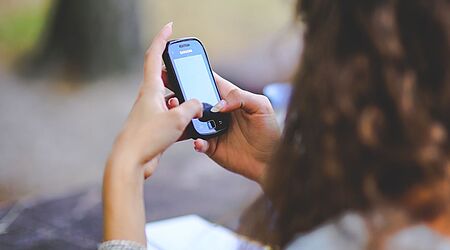 Der Hinterkopf einer jungen Frau mit langen lockigen Haaren ist zu sehen, die auf ein Handy schaut