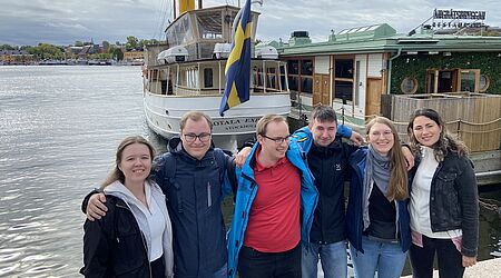 eine Gruppe junger Menschen steht, sich in den Armen haltend vor einer rustikalen Hafenkulisse mit Kutter