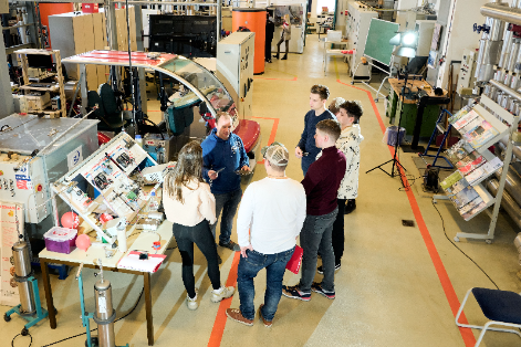 eine gruppe von jungen Menschen mit jemandem, der erklärt in einem großen Labor mit vielen technischen Aufbauten