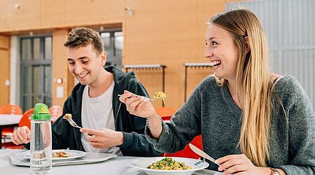 Zwei junge Menschen am Esstisch in der Mensa - lächelnd 