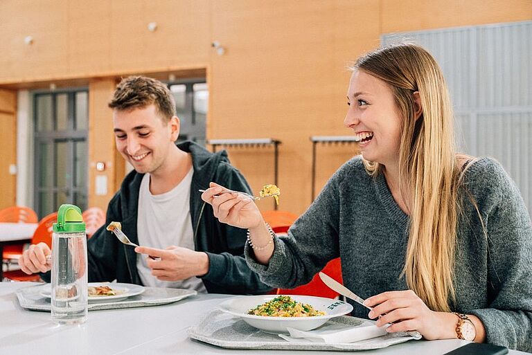 Zwei junge Menschen am Esstisch in der Mensa - lächelnd 