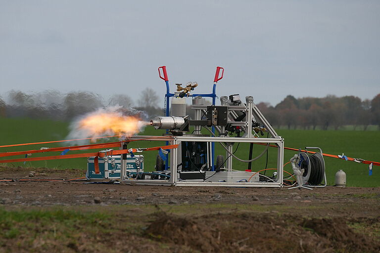 Ein Triebwerk, metallisch, mit Ausgangsrohren, liegt auf einem kleinen Traversensystem, eine dynamisch wirkende Flamme tritt aus