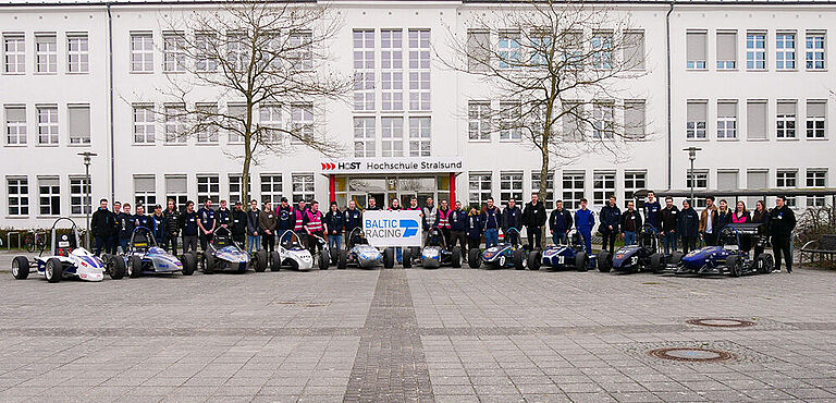 3 Dutzend Mitglieder des Baltic Racing Teams stehen mit 10 Generationen ihrer Formel-Student-Rennwagen vor der Fakultät für Elektrotechnik und Informatik