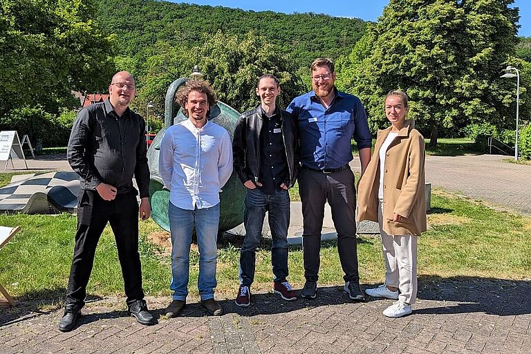 Fünf lächelnde Menschen - Das Team der HOST (v. l. n. r.): Martin Morgenstern, Torsten Wieck, Waldemar Adam, Jan Schmidt, Maria Freyer - stehen auf Asphalt im Grünen vor blauem Himmel