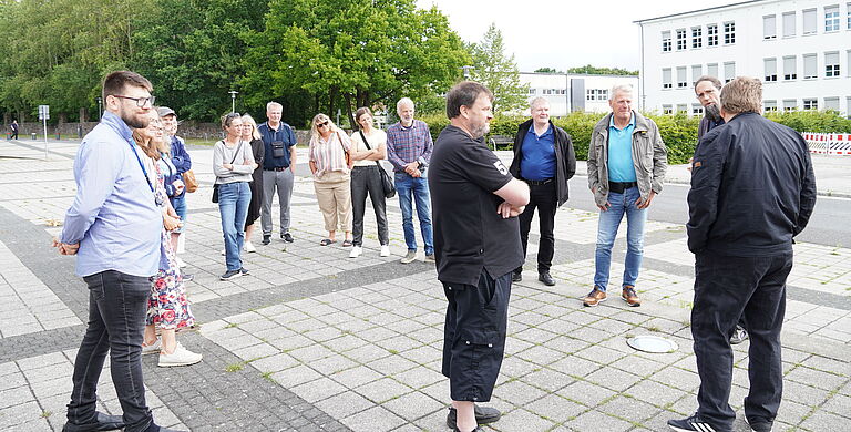 Eine Gruppe von Menschen steht auf dem Campusplatz, links vorn Daniel Hoffmann, und lauscht Prof. Dr. Johannes Gulden bei der Führung.