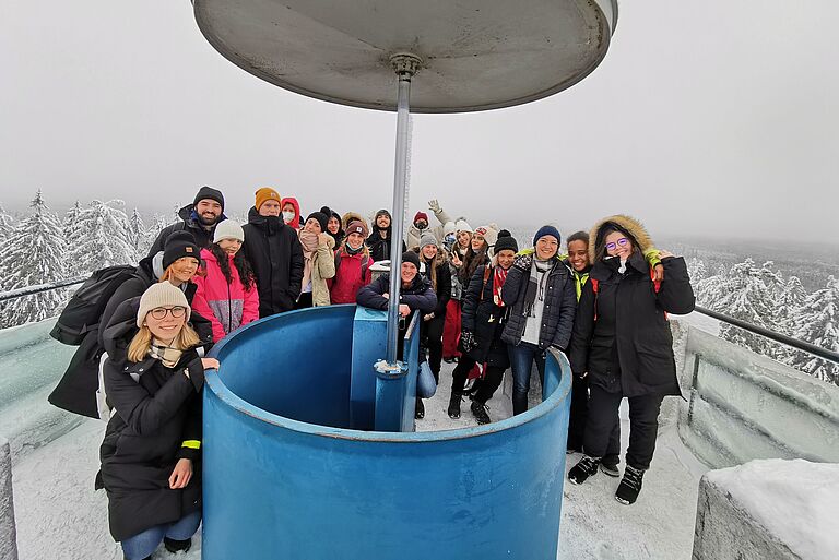 eine Gruppe junger Menschen steht auf einem Aussichtsturm in einer Winterlandschaft und lächelt von dort aus in die Kamera