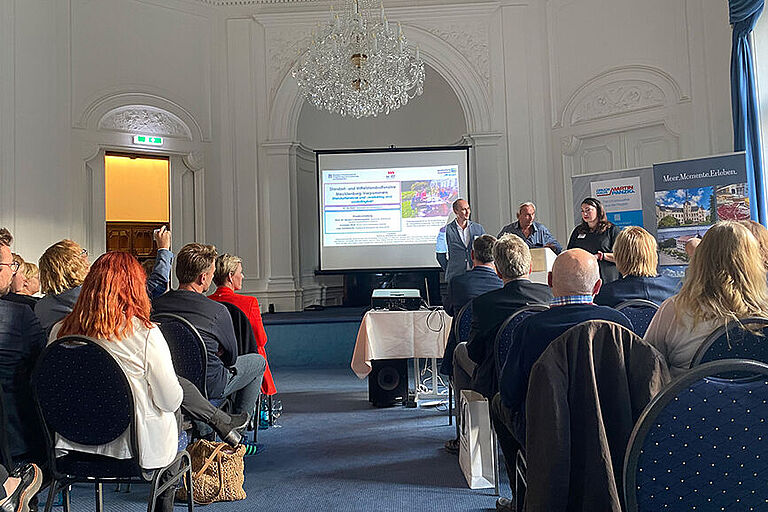 Mehrere Menschen sitzen in einem barock verzierten Saal bei einer Präsentation von Angehörigen der HOST.