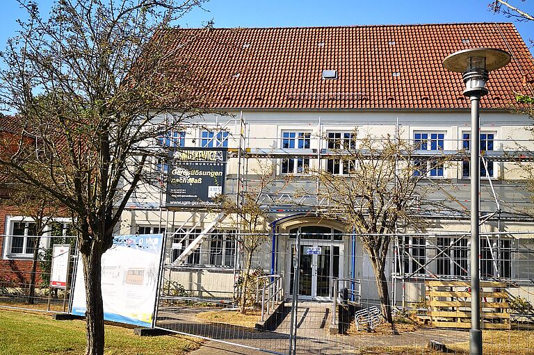 eine Ansicht der Bibliothek, ein weißes Gebäude, davor ein Bauzaun und Bäume