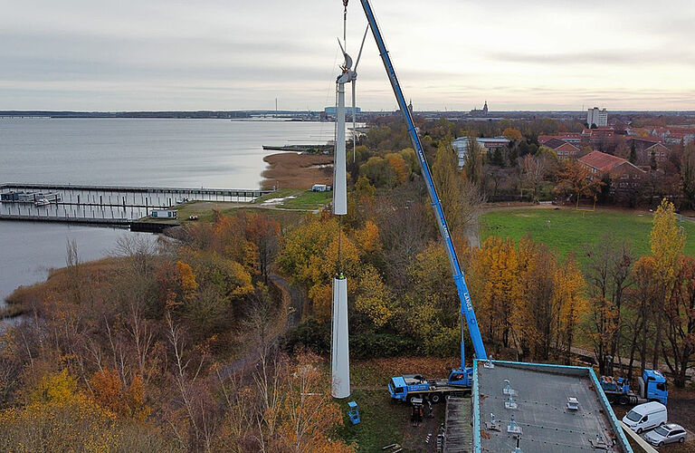 Luftbild der Kranmontage des 3-flügligen Windrads der Hochschule mit dem Strelasundim Hintergrund