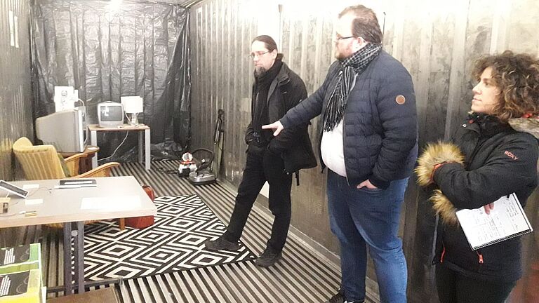 Prof. Dr. Johannes Gulden (l.) erhält einen Einblick in den Escape Room: Drei Personen stehen im Raum, vor ihnen Tisch, Stühle, ein Staubsauger, alte Computer, ein Fernseher, ein Koffer. 