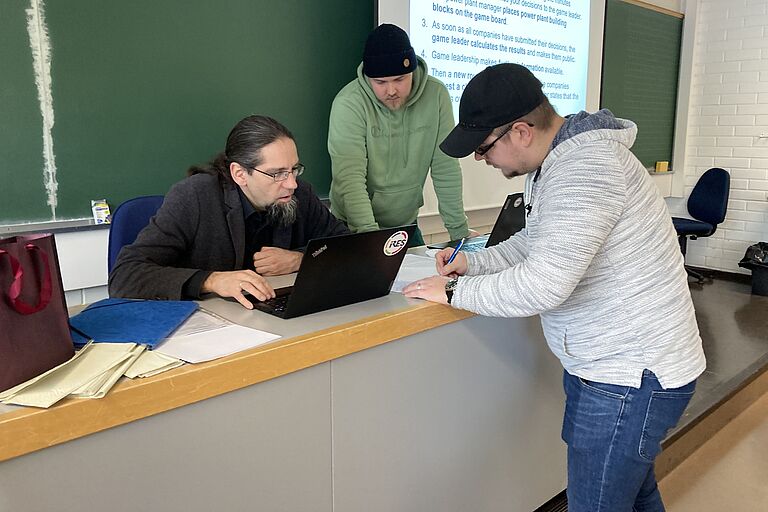 Ein Mann sitzt an einem Pult an einem Laptop, rechts neben ihm zwei jüngere Männer die mit auf Laptop oder Unterlagen schauen. 