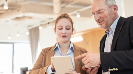 Eine junge Frau und ein älterer Mann schauen zusammen auf ein Tablet, beide tragen Blazer und stehen anscheinend in einem Büro.