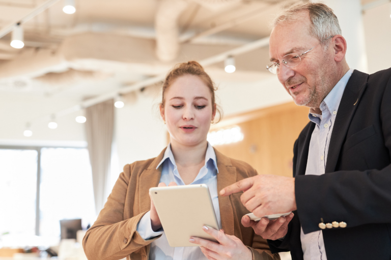 Eine junge Frau und ein älterer Mann schauen zusammen auf ein Tablet, beide tragen Blazer und stehen anscheinend in einem Büro.