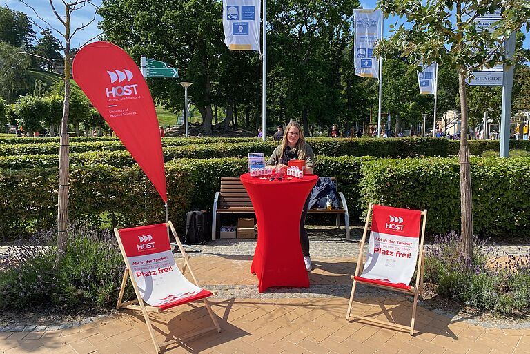 Eine junge Frau steht an einem roten Stehtisch der Hochschule Stralsund vorn zwei Liegestühle, neben ihr eine Beach-Flag - alles in Rot-Weiß und mit HOST-Logos.