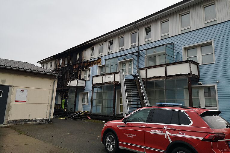 Ein feuerwehrauto steht vor einem Flachbau mit Holzfassade, Brandspuren sind erkennbar.