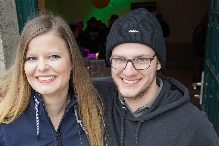 Paul Kunze und Anja Wachter vom AStA stehen lächelnd vor der Tür zum Haus 8, in dem bunte Lichter Feierstimmung verkünden und Silhouetten von Menschen zu erkennen sind