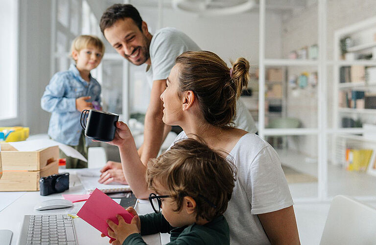 A family with two children is sitting at a table in a bright, light-flooded room. The parents are drinking coffee and smiling at each other.