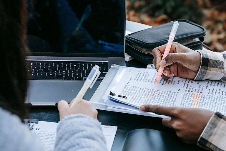 ein Symbolbild von einem Laptop und Händen von Studierenden mit Stift