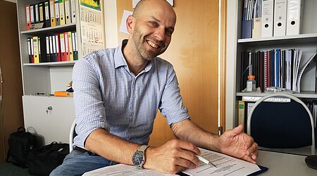 ein lächelnder Mann mit Unterlagen vor sich und einem Stift an einem Tisch sitzend