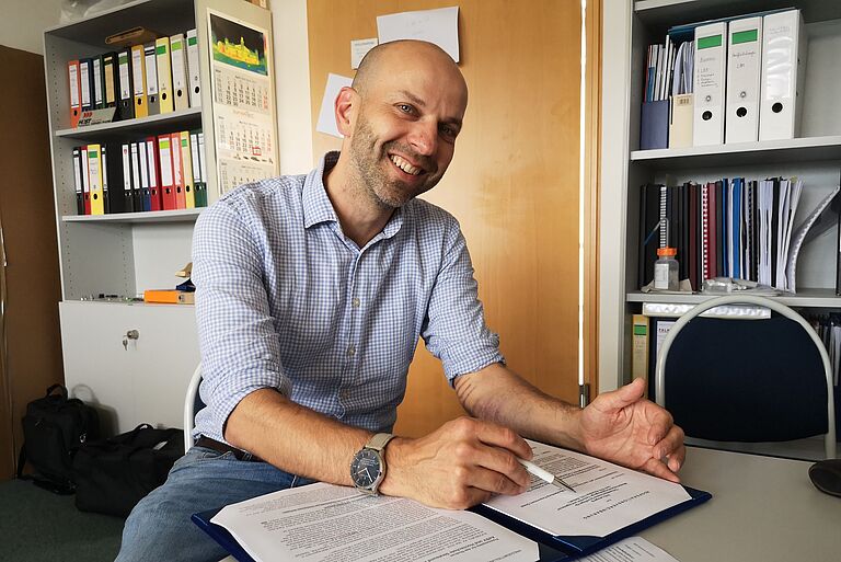 ein lächelnder Mann mit Unterlagen vor sich und einem Stift an einem Tisch sitzend