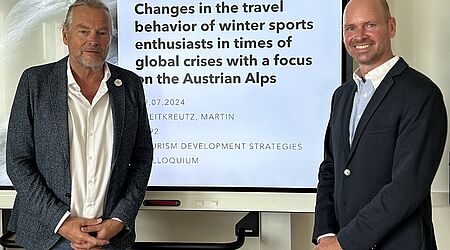 Zwei Männer stehen (leicht) lächelnd vor einem Bildschirm auf dem auf Englisch der Titel einer Abschlussarbeit zu lesen ist. "Changes in the travel behavior oft winter sports enthusiasts in times of gloabl crisis with a focus of the Austrian Alps"