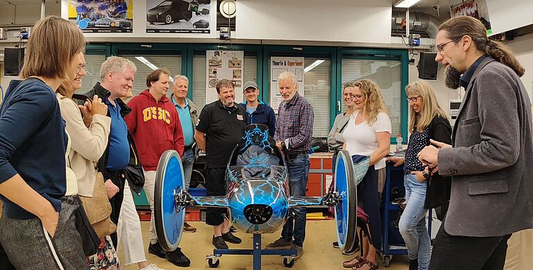 Eine Gruppe von Menschen, die Lehrenden aus Skandinavien umringt einen schlanken aufgebockten Rennwagen (den ThaiGER des Instiutes) - rechts steht der Leiter Prof. Dr. Johannes Gulden und erklärt offensichtlich etwas