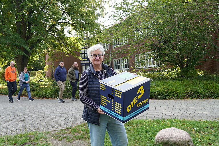 Eine Frau steht mit einer Umzugskiste vor einem Gebäude - die Leiterin vor Bibliothek.