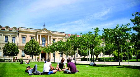 Ein großes gebäude, eine uni, vor der auf einer grünen Wiese junge Menschen sitzen.