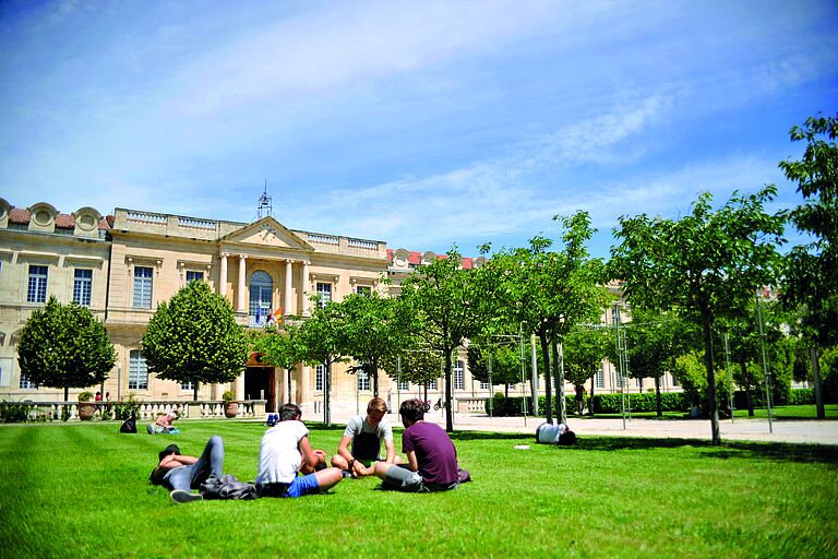 Ein großes gebäude, eine uni, vor der auf einer grünen Wiese junge Menschen sitzen.