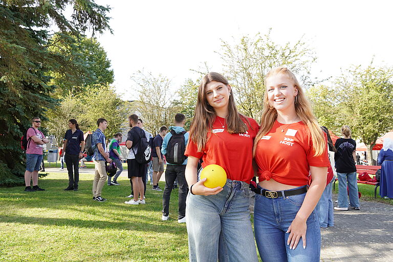 Zwei junge Frauen stehen in roten T-Shirts vor einer Gruppe junger Menschen, den neuen Studierenden