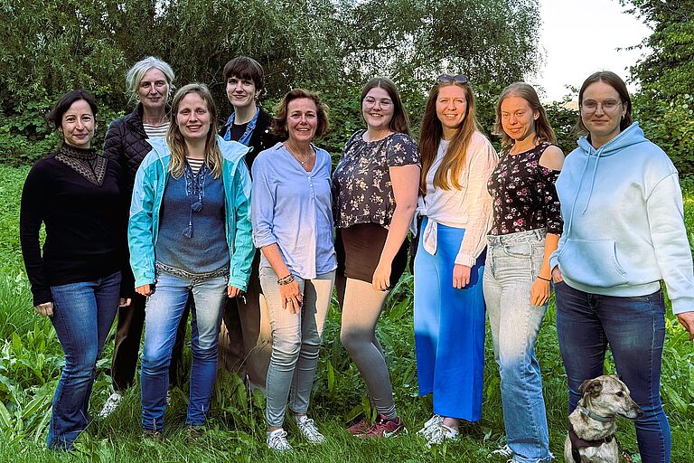 Eine Gruppe von Frauen verschiedener Alter, aber vorwiegend im Studierendenalter stehen lächeln im Grünen zusammen, vorn rechts ein kleiner Hund.