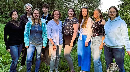 Eine Gruppe von Frauen verschiedener Alter, aber vorwiegend im Studierendenalter stehen lächeln im Grünen zusammen, vorn rechts ein kleiner Hund.