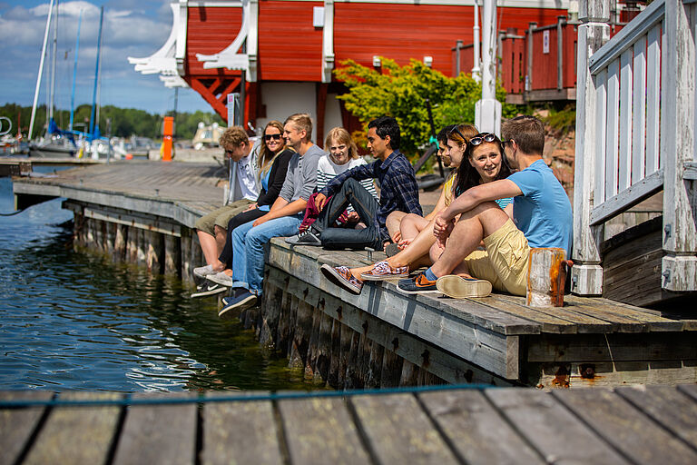 eine Gruppe junger, fröhlicher Menschen sitzt, sommerlich gekleidet, auf einem Steg.