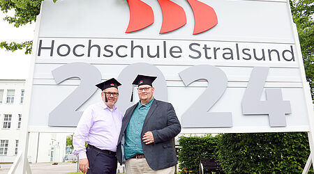 Zwei Männer mit Absolventenkappen stehen vor einem Hochschul-Schild mit einer großen 2024 darauf. 