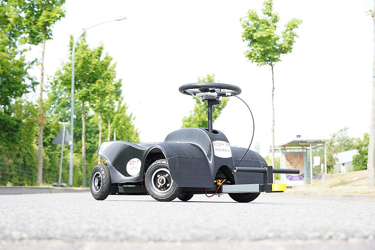 Das schwarze Rutschauto für Erwachsene steht auf der Straße, im Hintergrund sind Bäume zu sehen