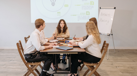 Einige junge Menschen sitzen an einem vermeintlichen Besprechungstisch vor einer Beamerprojektion.